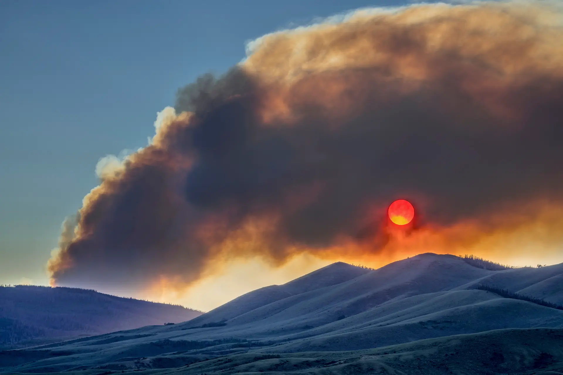 A wildfire smoke column over ridgelines at sunset, the sun deep red behind it.